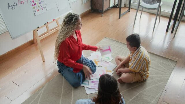 High angle view of female Latin tutor holding visual cards and demonstrating correct pronunciation of letters at speech therapy session with two diverse kids