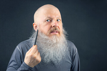 portrait of a bald man combing his large gray beard on a gray background.