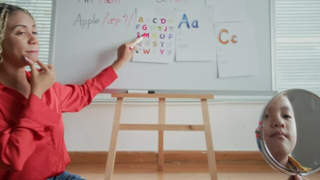 Handheld shot of interactive speech therapy class when female tutor pointing at letters on whiteboard and asking Asian schoolboy to repeat sounds correctly after her using mirror