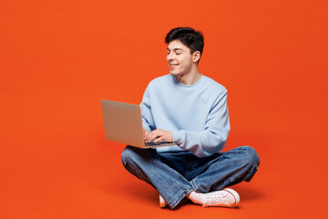 Full body young smart Caucasian IT man he wears blue sweatshirt casual clothes sits hold use work on laptop pc computer isolated on plain red orange color background studio portrait Lifestyle concept