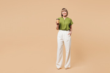 Fototapeta premium Full body elderly blonde woman 50s years old she wear green shirt casual clothes hold takeaway delivery craft paper brown cup coffee to go isolated on plain pastel beige background. Lifestyle concept.