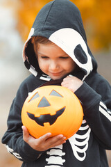 Cute and happy little boy in halloween skeleton costume holding a pumpkin bucket full of candies. Closeup child in spooky wear. 