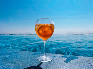 Photo of refreshing aperol cocktail in a wine glass, set against a stunning backdrop of  blue pool under a bright sky. The glass, perched on the edge of the pool, has condensation and an orange slice.