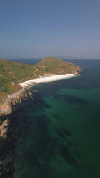 Soaring over Ons Island's turquoise waters on a sunny day, gliding forward toward the pristine Melide Beach. Lush green landscape with rocks and white sandy shores contrast against the deep blue sea.
