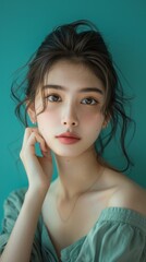 Fototapeta premium Confident Asian Woman in Portrait with Hand Touching Face on Teal Background in Natural Light
