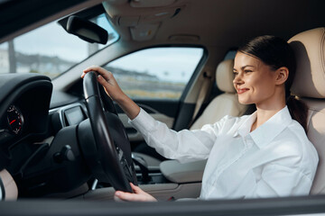 A smiling woman is seated in the driver's seat of a car, her hand gently resting on the steering...