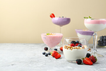 Different tasty yogurts with fresh berries and granola in glass dishware on gray textured table against pale yellow background, space for text