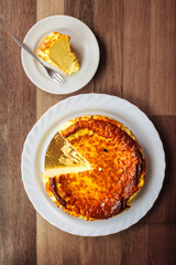 Famous Basque cheesecake from San Sebastian, Spain, on wooden table, served on white plates. Whole cake with portion removed served on smaller plate, showing a Rich, creamy texture.