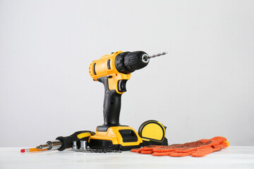 Cordless electric drill, gloves, bits, screwdriver and measuring tape on white wooden table