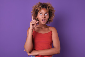 A young girl stands confidently with one finger raised in a moment of inspiration. Her curly hair and bright red top contrast beautifully against the vivid purple background.