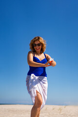 Sun protection. Beautiful elegant woman with long hair in blue swimsuit and white sarong and sunglasses applying sunscreen to her arms standing on white sandy beach on sunny day. Healthy lifestyle.