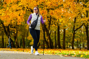 Smiling middle-aged blonde woman dressed sporty walking with poles on background of beautiful autumnal trees. Front view. Autumnal training of Nordic walking.