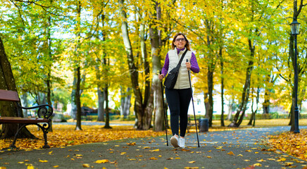 Fototapeta premium Beautiful middle-aged blonde woman dressed sporty exercising nordic walking on alley full of yellow fallen leaves in park on autumn day. Front view. Autumnal training of Nordic walking.