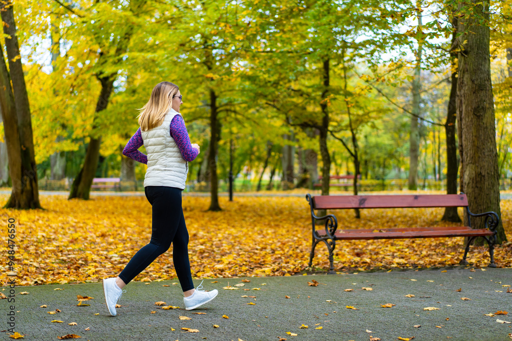 Wall mural Pretty middle-aged blonde woman dressed sporty running on alley full of beautiful autumnal trees in city park on autumn day. Back view. Autumnal training. Healthy running.