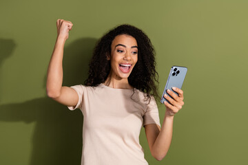 Photo of overjoyed girl with wavy hairdo dressed beige t-shirt hold smartphone raising fist win bet isolated on khaki color background