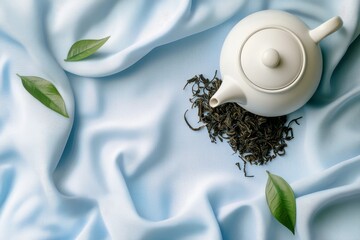 White teapot with loose leaf tea on blue fabric, complemented by green leaves, creating a calming and elegant tea time setting.