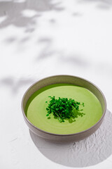 Green cream soup with microgreens in bowl standing on white table with shadows