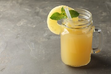 Tasty pineapple juice in mason jar, mint and slice of fruit on grey textured table, closeup. Space for text