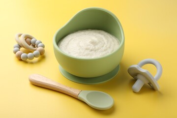 Delicious baby food in bowl with spoon, pacifier and teether on yellow background