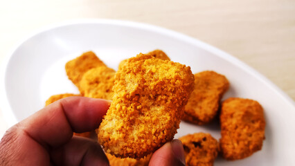 Fingers grabbing chicken breast fillet in a crispy breading known as Nuggets on a white plate.
