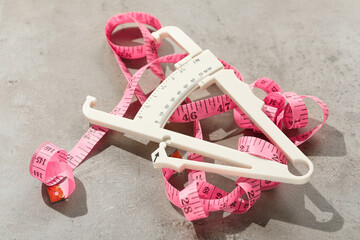 Plastic body fat caliper and measuring tape on grey table, closeup