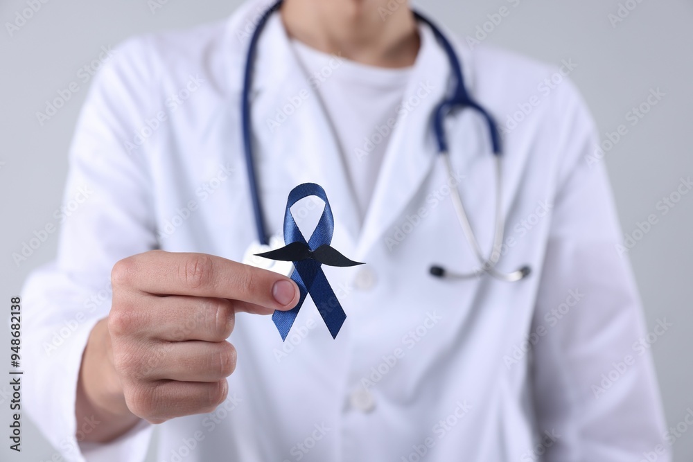 Wall mural Prostate cancer awareness. Doctor holding blue ribbon with fake mustache on light grey background, closeup