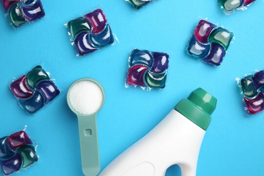 Bottle of fabric softener, laundry detergent pods and measuring scoop of washing powder on blue background, flat lay