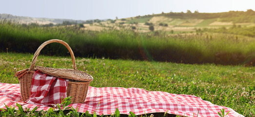 Picnic blanket and wicker basket on green grass during sunny day. Banner design with space for text