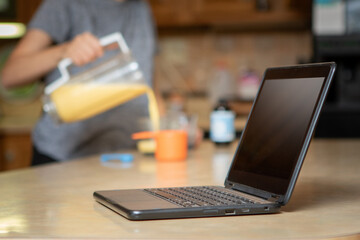 Young woman pouring drink and cooking while listening to audiobook on laptop in kitchen