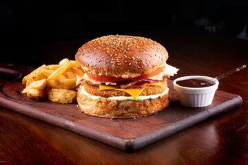 Breaded chicken burger with bacon, lettuce and tomato and potatoes from above on wooden board with bbq sauce black background
