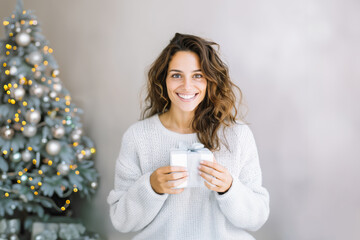 Portrait of a young Spanish woman smiling while holding a gift box next to a decorated Christmas tree.