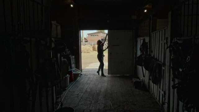 Wide shot of young female equestrian opening equine shed gates before horsemanship training, backlight. Outside barn gate there is serene rural outdoor setting with wooden sheds and haystacks