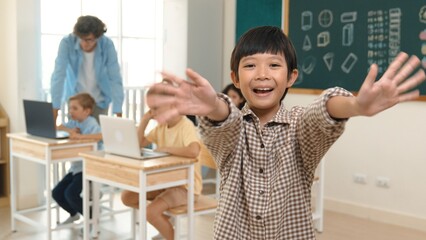 Asian boy looking at camera while waving hands and standing at blackboard. Skilled child greeting while diverse student study or learning about coding engineering prompt and generated AI. Pedagogy.