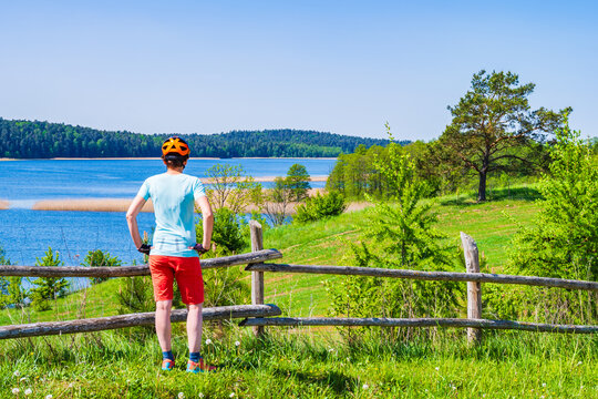 Woman cyclist tourist standing against wooden fence looking at Wigry lake and green landscape, Wigry National Park, Podlasie, Poland
