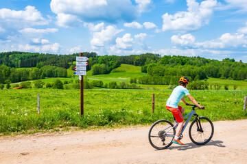 Woman cyclist riding bicycle on gravel rural road among green meadows, Suwalski Landscape Park,...