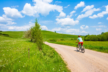 Fototapeta premium Woman cyclist riding bicycle on gravel rural road among green meadows, Suwalski Landscape Park, Podlasie, Poland
