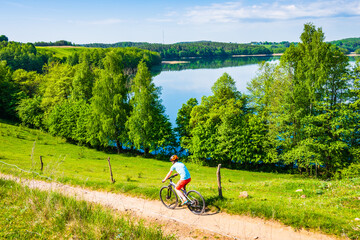 Woman cyclist riding bicycle on gravel rural road along lake and among green meadows, Suwalski...
