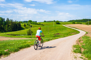 Obraz premium Woman cyclist riding bicycle on gravel rural road among green meadows and fields, Suwalski Landscape Park, Podlasie, Poland