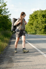 Young woman in casualwear and headband looking back at highway while carrying backpack and rolled mat during hitchhiking trip