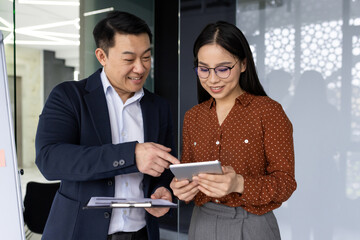Asian business professionals interacting in office environment, engaging with digital tablet. Smiling, discussing ideas, technology, collaboration. Emphasizing teamwork, modern workplace dynamics.
