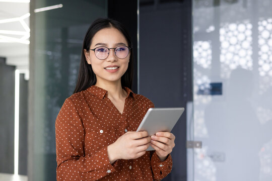 Confident Asian worker in modern office environment holding tablet. Dressed in polka dot blouse , she exhibits friendly and professional demeanor, representing diverse workforce in corporate setting - Powered by Adobe