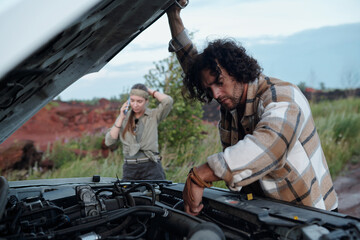 Young man with dark wavy hair bending over open hood of pickup truck and checking engine against his girlfriend calling repair service © pressmaster