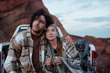 Young romantic couple sitting in body of pickup truck and looking aside while guy embracing his girlfriend holding cup of tea