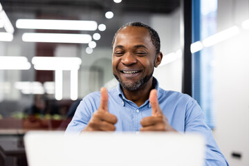 Happy businessman gives thumbs up during video call in office, showing approval and success. Confident and engaged professional enjoying virtual meeting or online conference interaction.