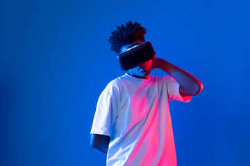 Surprised young African American looking through VR holding interesting object hologram on pink blue neon wall at metaverse world connecting digital futuristic technology virtual reality. Contrivance.