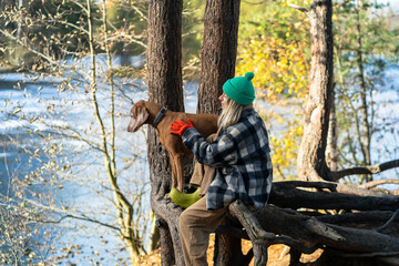 Joyful woman hiking with dog on cold autumn day sitting on tree roots near frozen river in woodland. Pleased female owner of Magyar Vizsla enjoys strolls with pet in golden forest park in late fall.