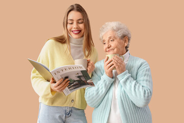 Young woman and her grandmother with cups of tea reading magazine on beige background