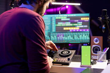 Music producer uses controls and knobs on mixing console in home studio, having a smartphone with greenscreen display. Artist learning to edit tunes and add sound effects with daw software.