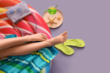 Young woman sitting on inflatable mattress near flip-flops and cocktail against purple background