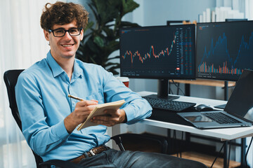 Profile of smiling young stock trader looking at camera, holding notepad and pen against on...